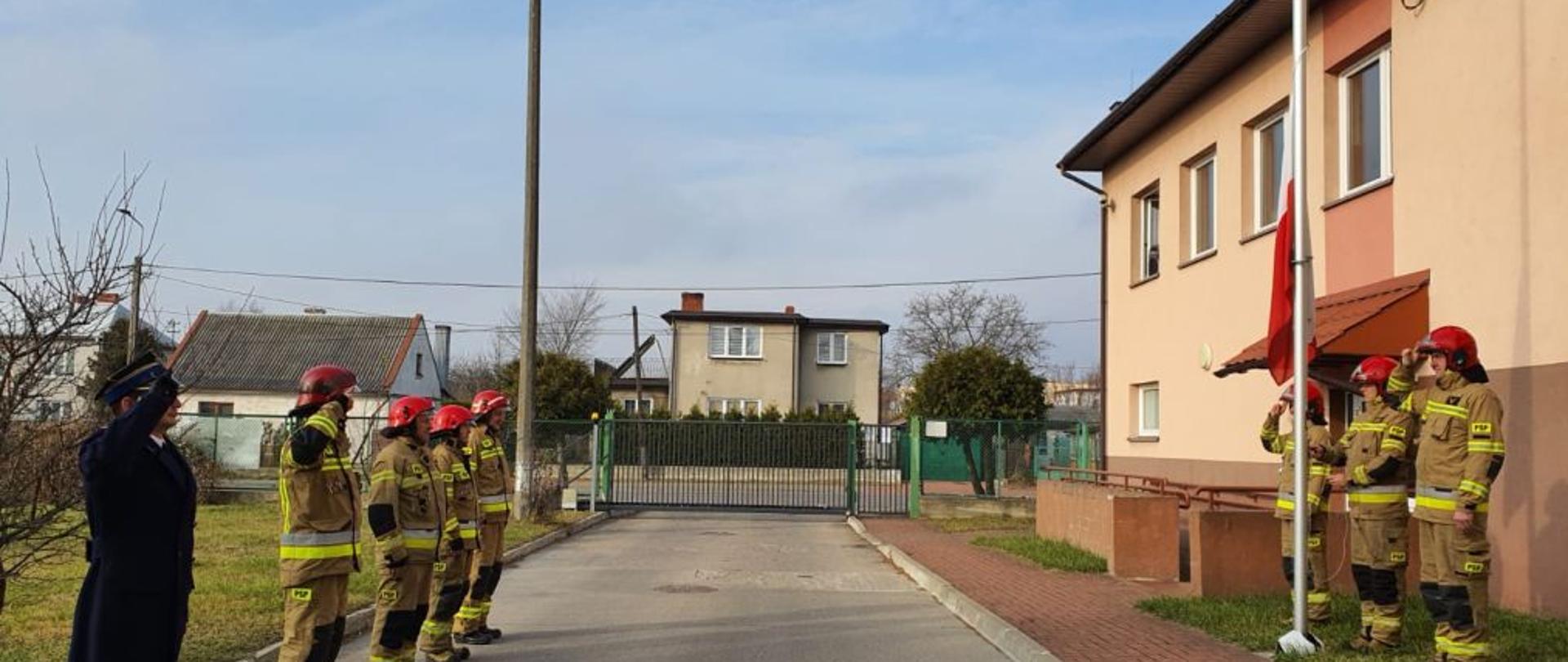 Przy maszcie flagowym stoi 3 strażaków w żółtych mundurach, jeden z nich podnosi flagę. Po drugiej stronie stoi 5 strażaków, 4 w żółtych mundurach jeden w granatowym płaszczu.