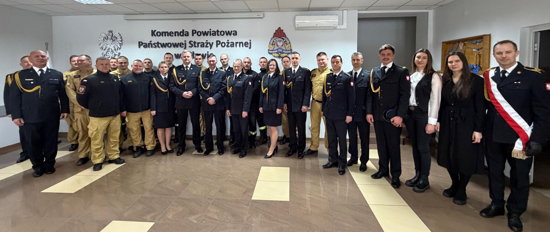 Zdjęcie grupowe wykonane w sali komendy. Funkcjonariusze PSP pozują do wspólnej fotografii. W tle widoczny jest napis „Komenda Powiatowa Państwowej Straży Pożarnej”, godło państwowe oraz emblemat straży pożarnej.