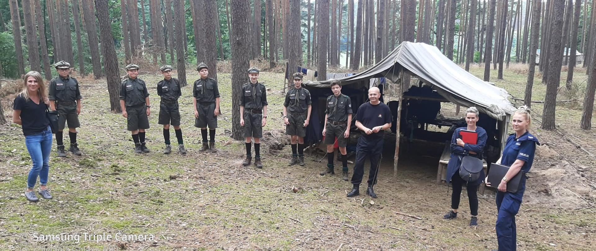 Zdjęcie przedstawia wypoczywających na leśnym obozie harcerzy wraz z przedstawicielami służb kontrolujących warunki wypoczynku oraz bezpieczeństwo młodzieży.