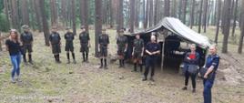 Zdjęcie przedstawia wypoczywających na leśnym obozie harcerzy wraz z przedstawicielami służb kontrolujących warunki wypoczynku oraz bezpieczeństwo młodzieży.