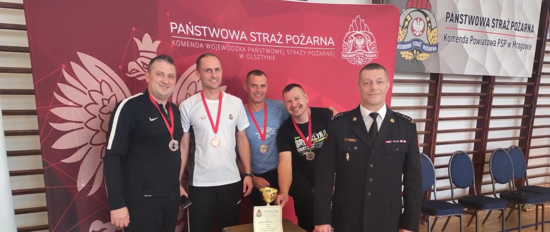 Na zdjęciu przy podium drużyna strażaków z Braniewa z Komendantem wojewódzkim. W tle baner czerwony Państwowa Straż Pożarna.