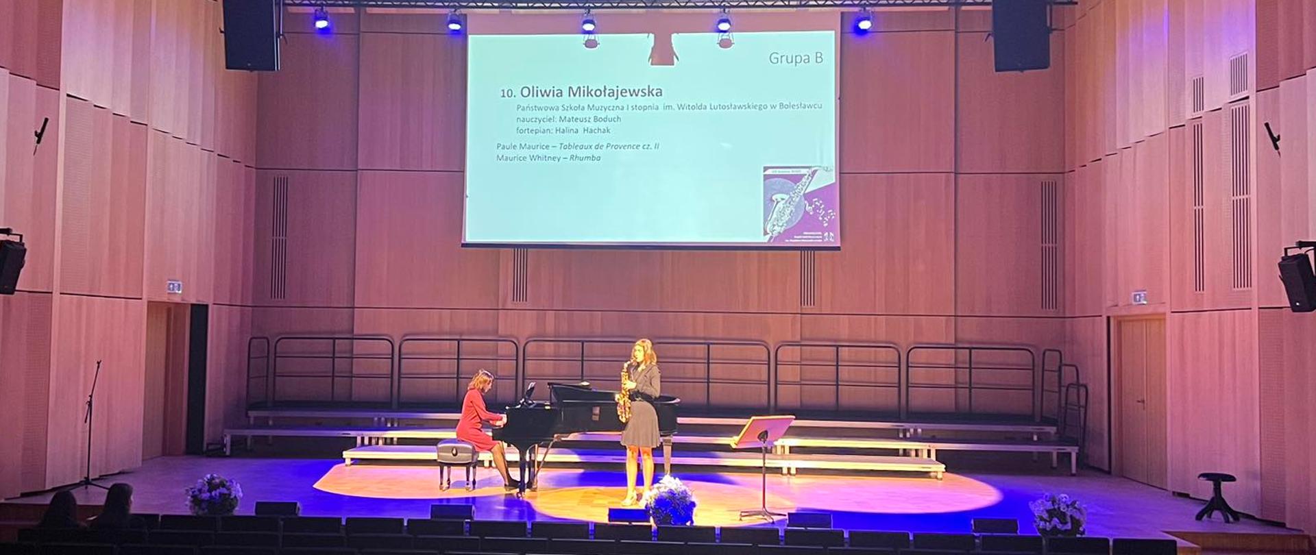 Zdjęcie przedstawiające w sali koncertowej, na scenie: Oliwię Mikołajewską (na saksofonie) i panią Hałynę Hachak (akompaniament fortepianowy), podczas II Ogólnopolskiego Konkursu Saksofonowego uczniów Szkół Muzycznych I i II stopnia w Zespole Szkół Muzycznych w Łodzi.