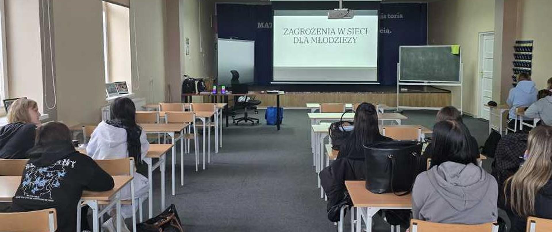 Zdjęcie przedstawia jasną, szkolną salę lekcyjną lub aulę widzianą z perspektywy tyłu klasy. W ławkach siedzą uczniowie, a ich uwaga skierowana jest na ekran projekcyjny znajdujący się na końcu pomieszczenia.Układ sali i ludziePierwszy plan i środek: Przez środek sali biegnie szerokie przejście wyłożone ciemnoszarą wykładziną. Po obu stronach przejścia ustawione są rzędy jasnych, drewnianych biurek i krzeseł. W ławkach siedzi kilkanaście osób, głównie młodzież (widoczna od tyłu), ubrana w codzienne stroje (bluzy, kurtki). Niektórzy mają przy ławkach plecaki i torebki.Tło (przód sali): Na końcu sali znajduje się podwyższenie (scena). Po prawej stronie stoi tradycyjna, zielona tablica szkolna na stojaku. Po lewej stronie widać biurko nauczyciela z krzesłem obrotowym oraz mniejszą, białą tablicę.Ekran projekcyjnyW centralnym punkcie z przodu sali wisi duży ekran. Wyświetlany jest na nim slajd tytułowy prezentacji. Slajd ma jasne tło z zielonymi paskami na górze i na dole. Na środku widnieje duży, czarny napis w języku polskim: „ZAGROŻENIA W SIECI DLA MŁODZIEŻY”.Architektura i oświetleniePomieszczenie ma wysoki, biały sufit z podłużnymi lampami. Po lewej stronie znajdują się duże, wysokie okna z opuszczonymi częściowo beżowymi roletami, które wpuszczają do sali naturalne światło. Ściany mają jasny, beżowo-szary kolor.