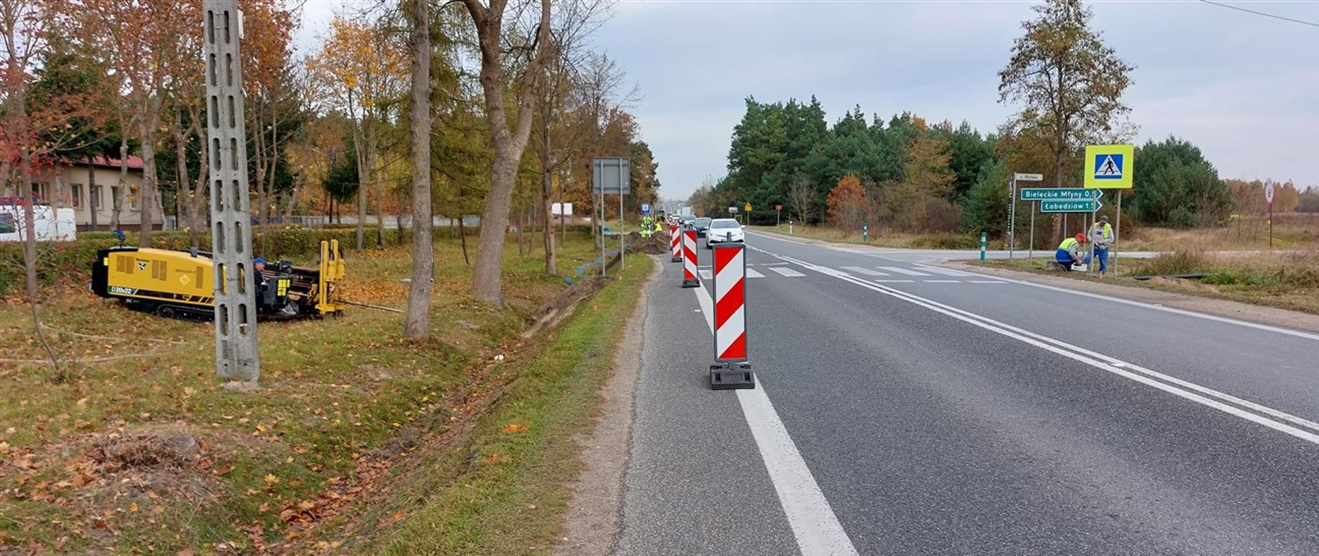 Droga krajowa nr 73 - pierwsze prace przy doświetlaniu przejść dla pieszych. Jednojezdniowa droga z zawężonym poboczem za pomocą biało-czerwonych elementów oznakowania. Przy drodze żółta maszyna do wykonywania kanałów pod przyłącza, w oddali ekipy budowlane po obu stronach drogi.