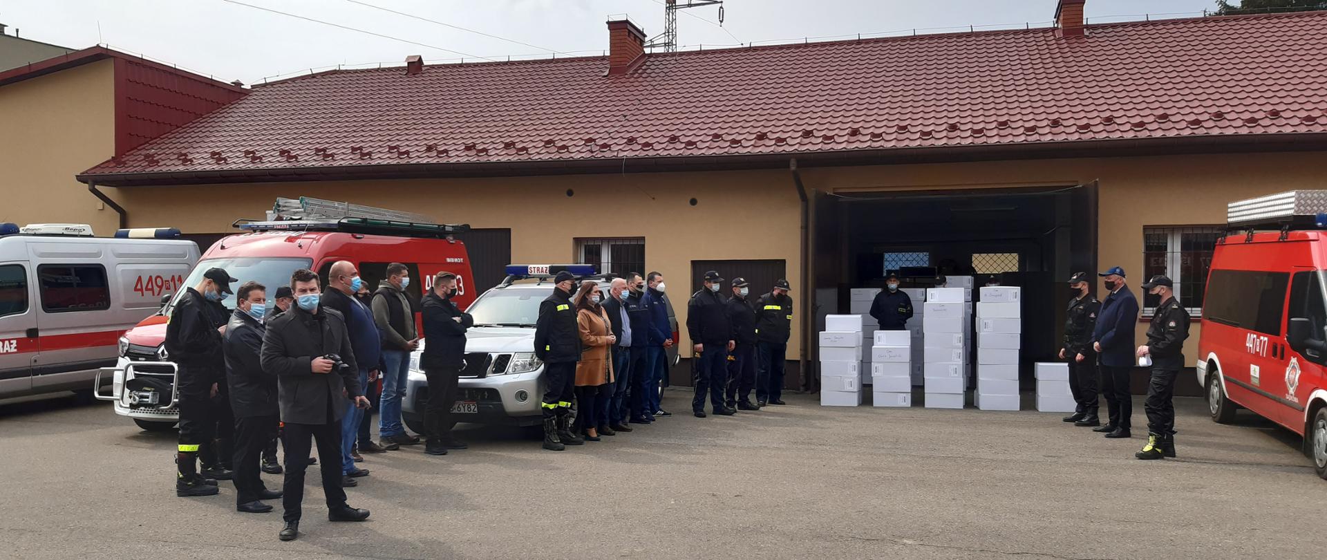 Na zdjęciu na placu wewnętrznym stoją komendant powiatowy państwowej straży pożarnej w Jaśle, starosta jasielski, zastępca komendanta powiatowego państwowej straży pożarnej w Jaśle oraz przedstawiciele z urzędów miast i gmin powiatu jasielskiego, przed garażem pudła z maseczkami chirurgicznymi