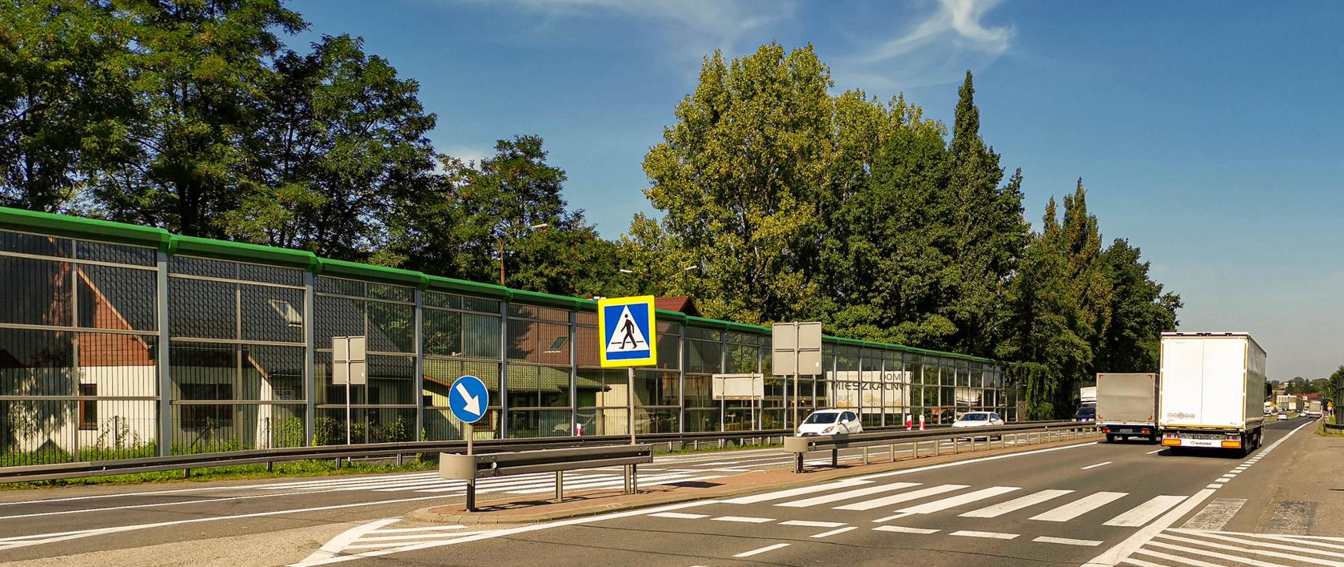 Fotografia z poziomu terenu na przejście dla pieszych przez DK1 w rejonie skrzyżowania z ul. Stawową w Goczałkowicach-Zdroju. Na dwujezdniowej drodze widoczny duży ruchu pojazdów. W centralnej części zdjęcia zlokalizowane jest przejście dla pieszych z wydzielona wyspą separacyjną pomiędzy dwupasowymi jezdniami. Droga wyposażona w ekrany akustyczne. 