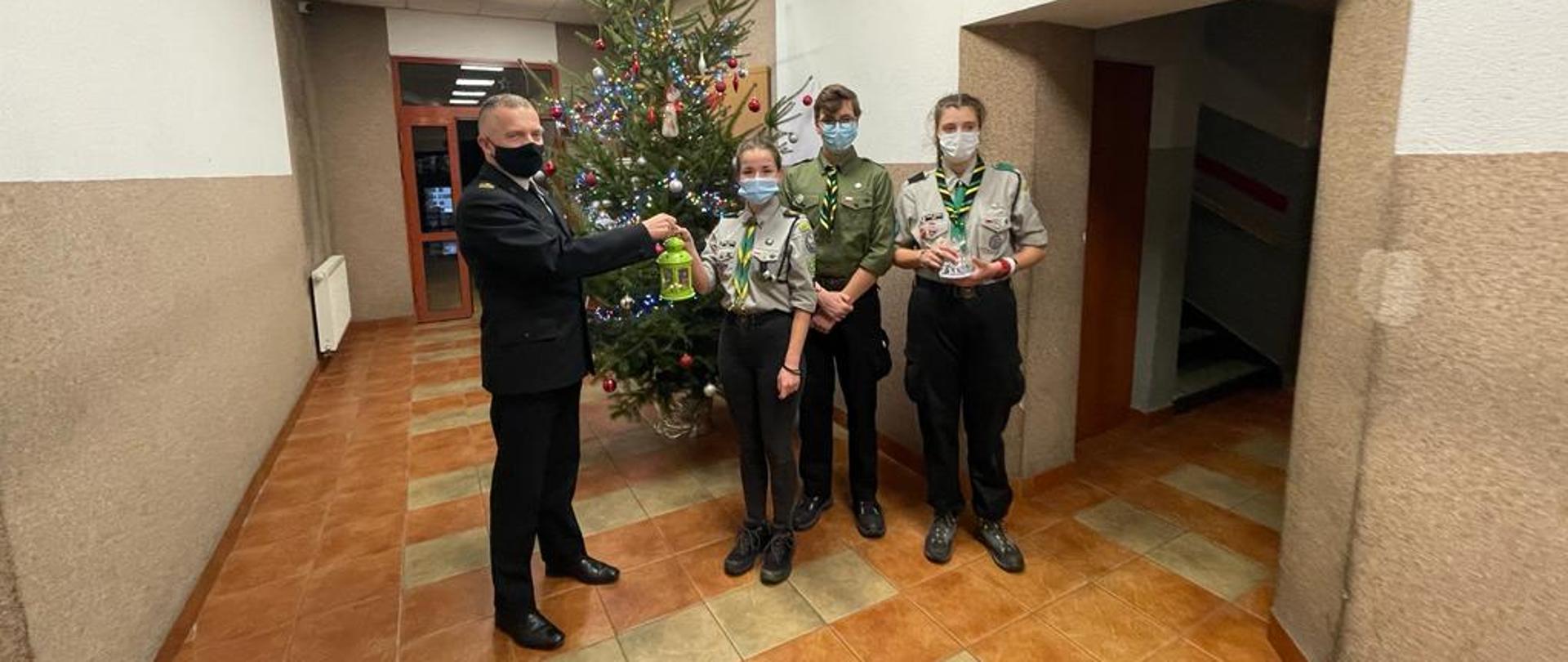 Zdjęcie przedstawia Komendanta Powiatowego Państwowej Straży Pożarnej w Będzinie wraz z harcerzami przekazującymi symboliczne "światło pokoju" na holu przy choince.