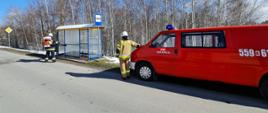 Widoczny od tyłu strażak w ubraniu specjalnym i hełmie strażackim dezynfekuje przystanek autobusowy przy użyciu przenośnego plecakowego opryskiwacza spalinowego. Obok, po prawej stronie, widoczny od tyłu obok samochodu pożarniczego w kolorze czerwonym, drugi strażak w ubraniu specjalnym.