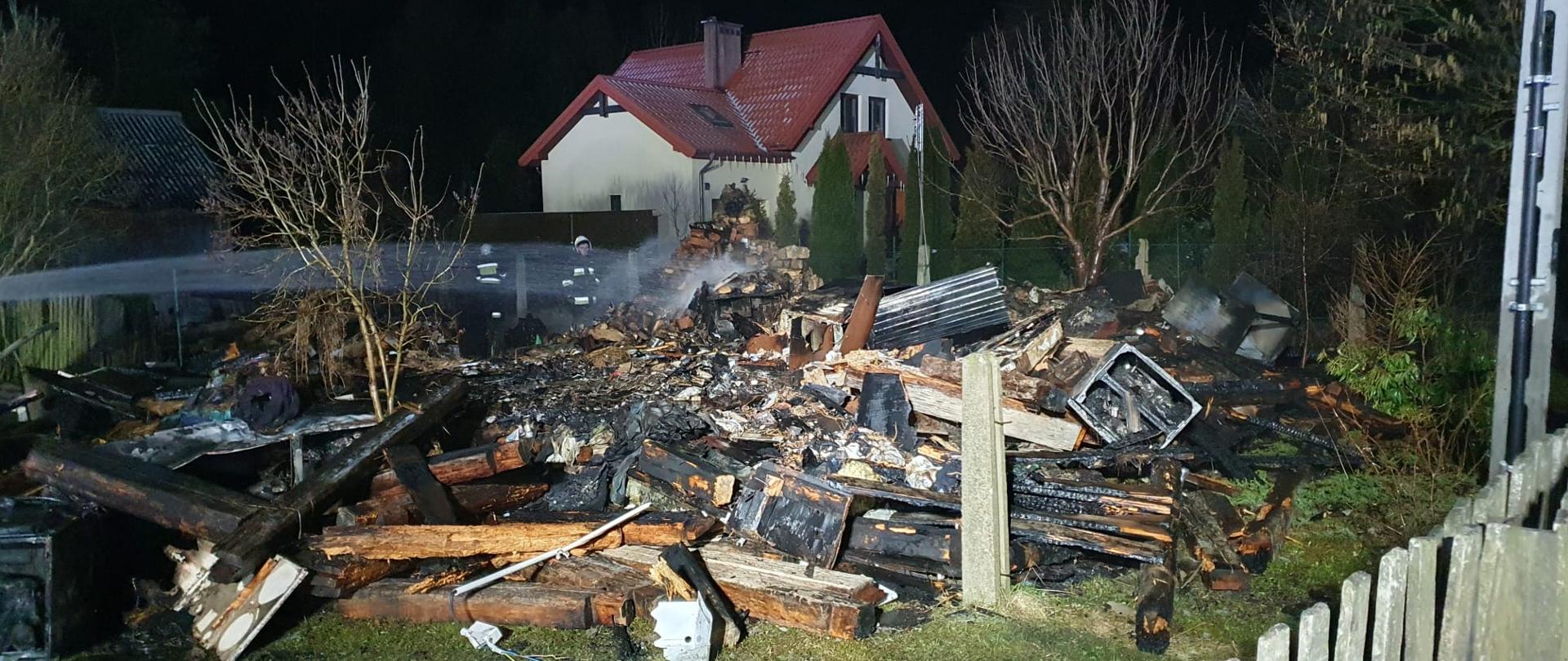 Zdjęcie przedstawia pogorzelisko pozostałe po pożarze budynku mieszkalnego. Z lewej strony widać strumień wody podawany w celu dogaszenia, a w tle sąsiadujący ze spalonym obiektem dom.
