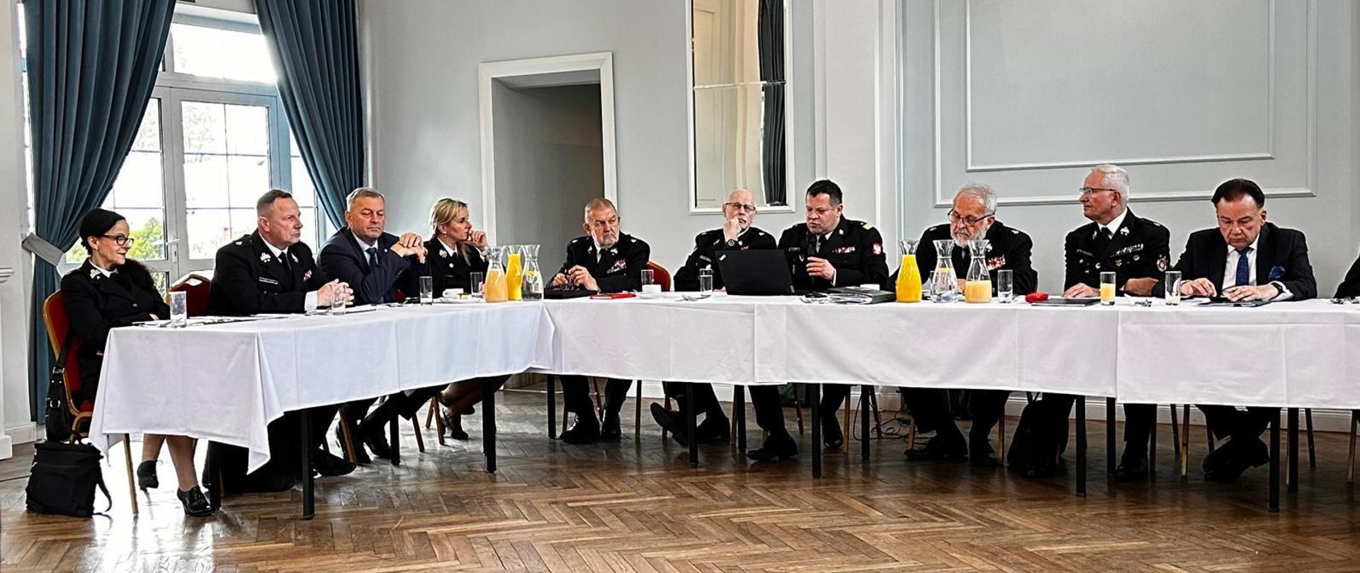 Do mikrofony mówi siedzący za stołem Mazowiecki Komendant Wojewódzki PSP nadbryg. Artur Gonera. Na zdjęciu widocznych kilkunastu innych gości siedzących za stołem. 