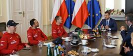 Minister Kamil Bortniczuk oraz grupa żużlowców w czerwonych reprezentacyjnych bluzach rozmawiają przy stole. Na stole znajdują się napoje, owoce, woda. W tle widoczne flagi Polski i Unii Europejskiej.