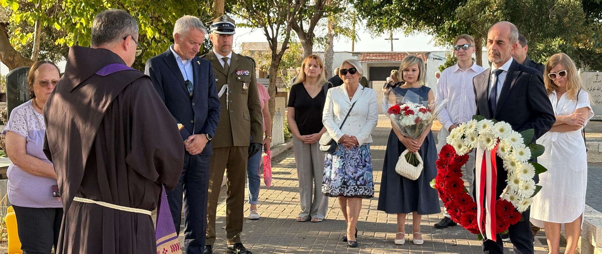 Grupa ludzi z refleksyjną mimiką twarzy, zamyśleni. Na pierwszym planie mężczyzna z biało czerwonym wieńcem kwiatowym. 