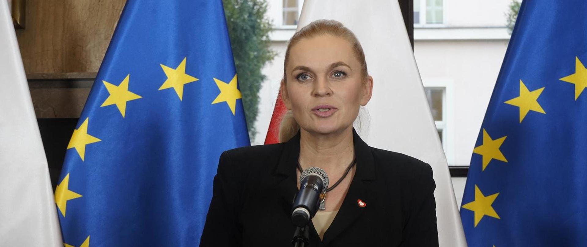 Minister Nowacka stoi i mówi do mikrofonu na stojaku, za nią pod ścianą rząd flag Polski i UE.
