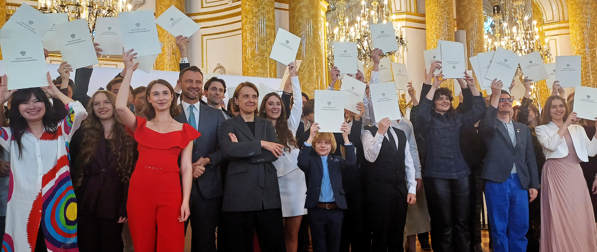 duża grupa osób w galowych strojach trzymających nad głowami dyplomy, w tle bogato zdobiona sala zamkowa