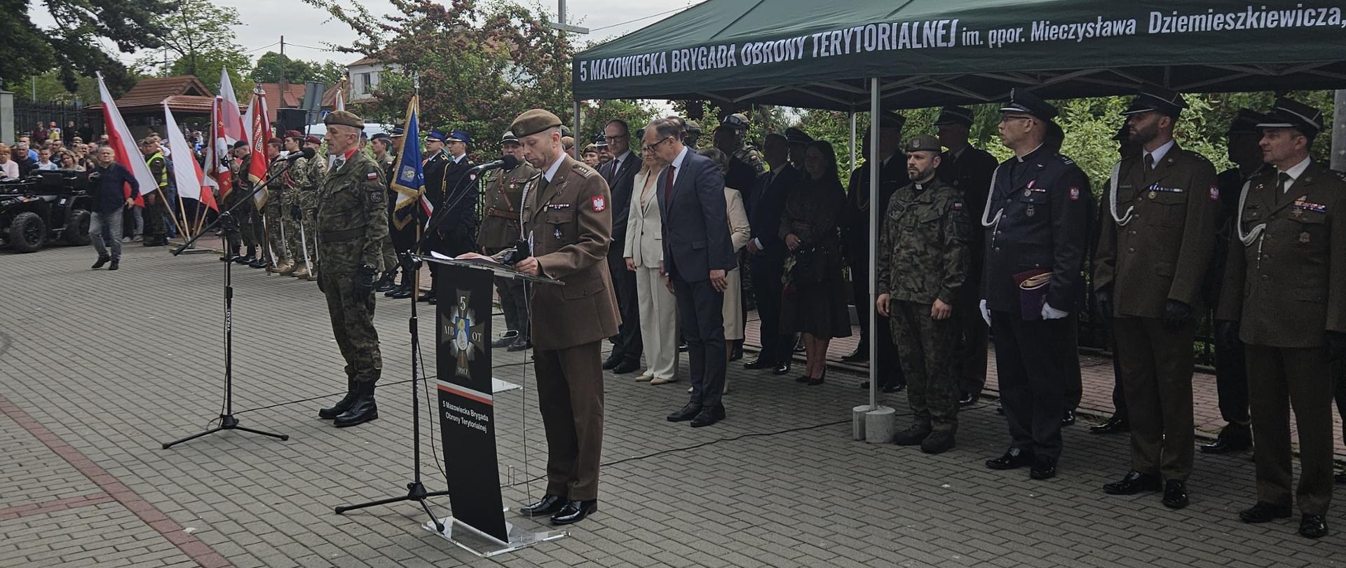 zdjęcie z uroczystości, grupa ludzi, uczestników wydarzeń stoi w rzędzie na drugim planie, na pierwszym planie przy mównicy stoi żołnierz