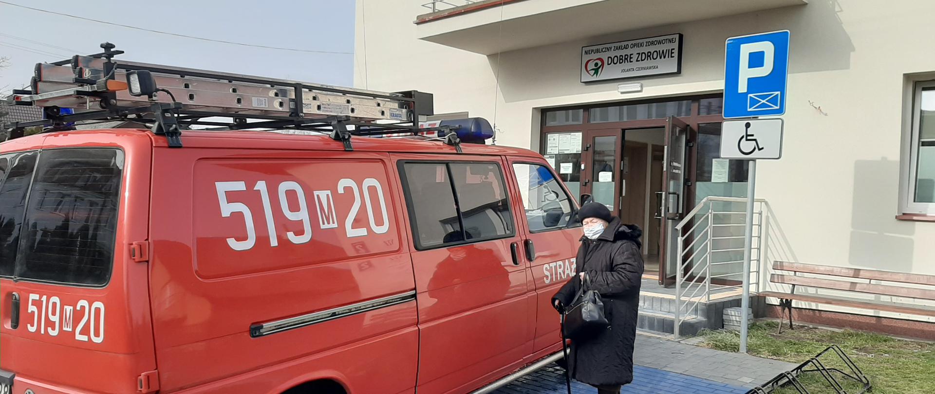 Starsza kobieta stojąca przy samochodzie jednostki OSP Rudzienko zaparkowanym przed przychodnią