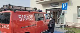 Starsza kobieta stojąca przy samochodzie jednostki OSP Rudzienko zaparkowanym przed przychodnią