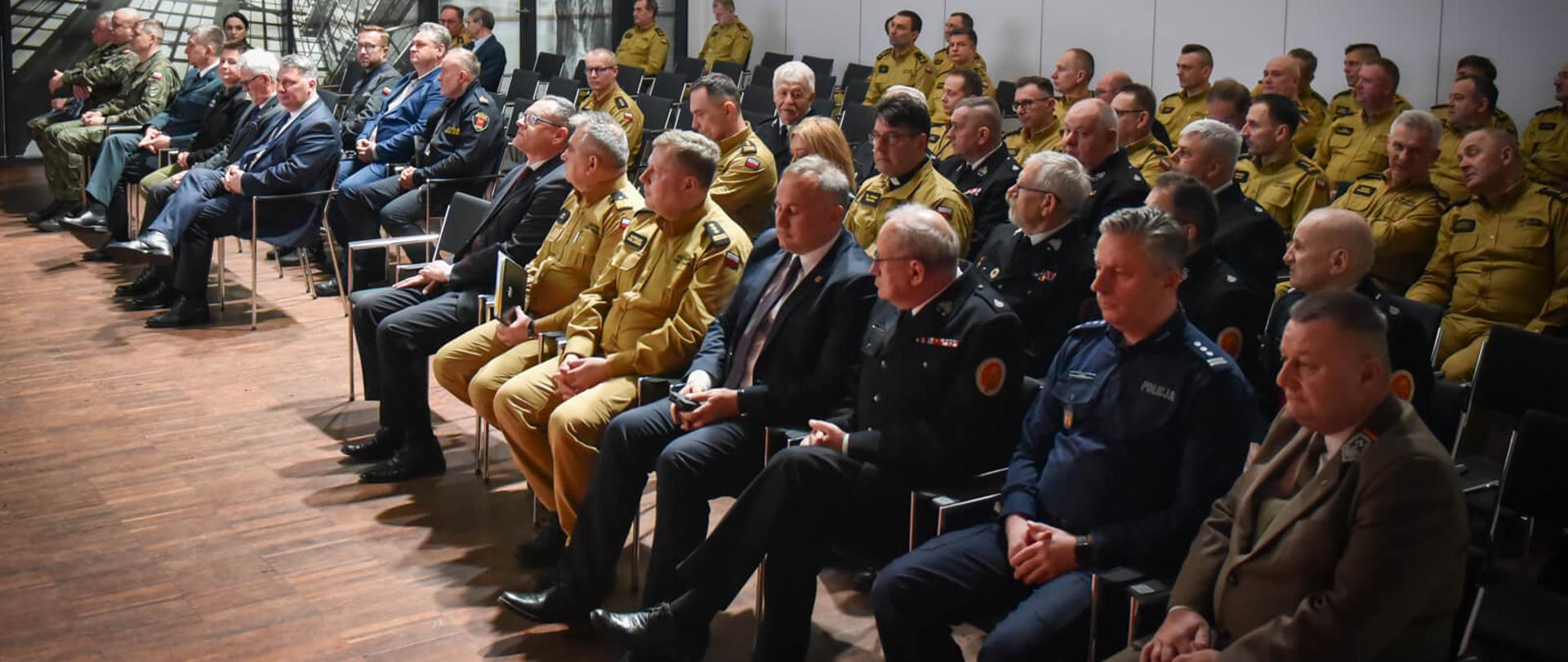 uczestnicy narady siedzący na krzesłach w sali konferencyjnej, część z nich to strażacy w umundurowaniu służbowym