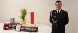 Na zdjęciu Pan asp. szt., z boku na stoliku okolicznościowe prezenty, które otrzymał tj.: ozdobny miecz umieszczony na ozdobnej desce, ryngraf oraz replika samochodu strażackiego Jelcz