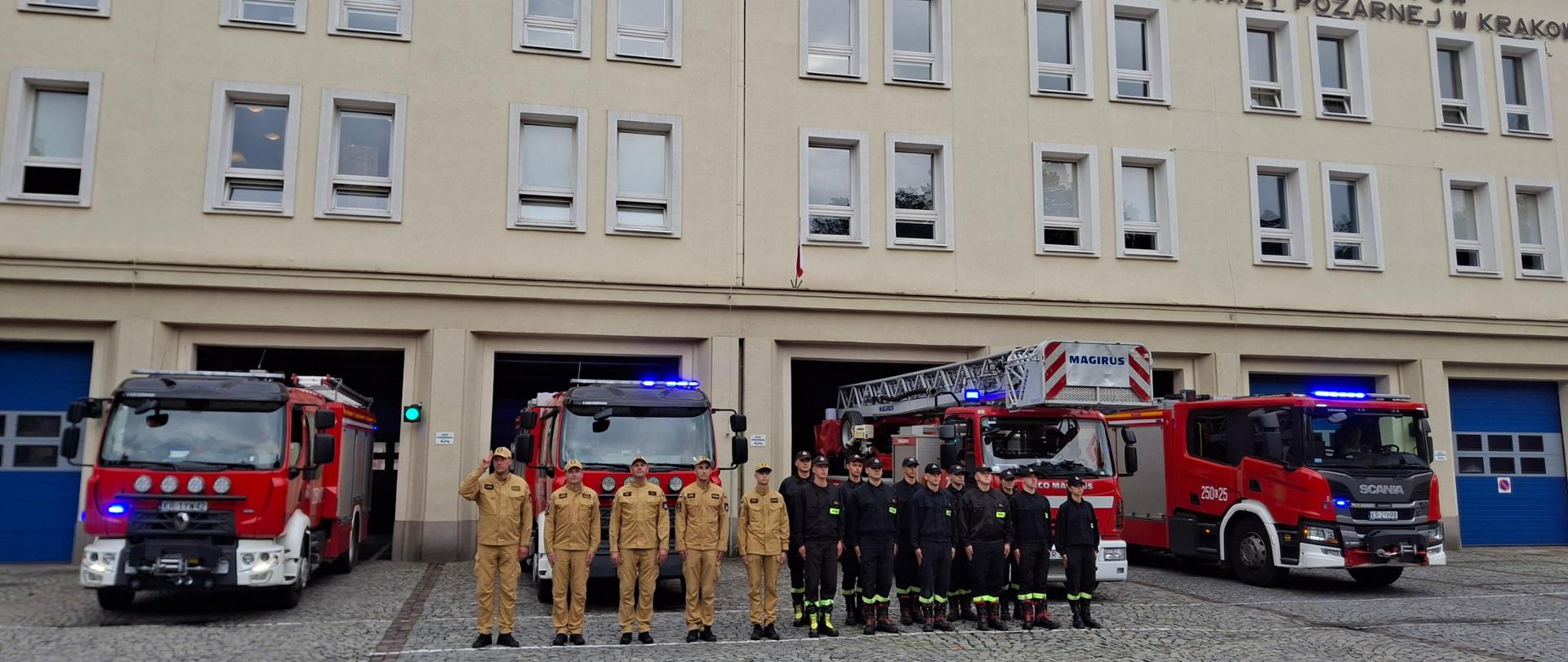 Plac zewnętrzny Szkoły Aspirantów PSP w Krakowie. Przed bramami garażowymi ustawione samochody ratowniczo-gaśnicze z włączonymi sygnałami świetlnymi, a przed nimi kadra podziału bojowego w pozycji zasadniczej z salutującym dowódcą. 