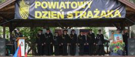 strażacy i druhowie stoją na drewnianej zadaszonej scenie, nad nimi baner z napisem powiatowy dzień strażaka