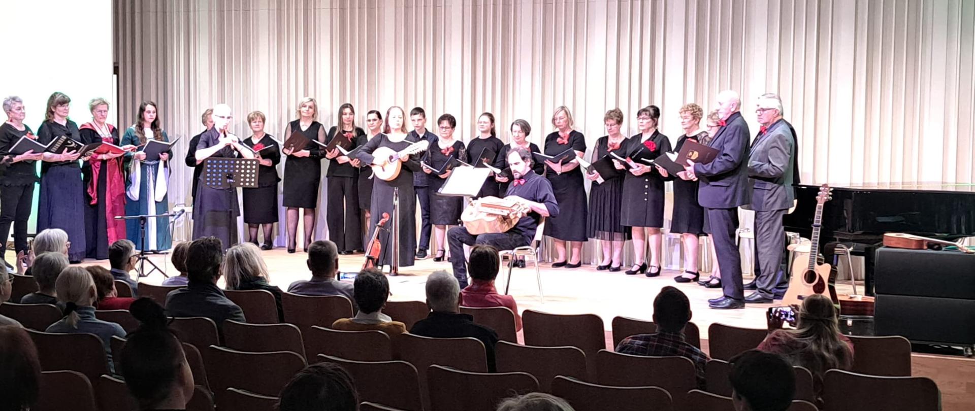 Na scenie duża grupa odświętnie ubranych starszych kobiet i mężczyzn. Stoją w półokręgu, a w środku dwie kobiety grające na instrumentach oraz siedzący na krześle mężczyzna. Tło obrazka w odcieniach beżu. Bokiem artystów czarny fortepian.