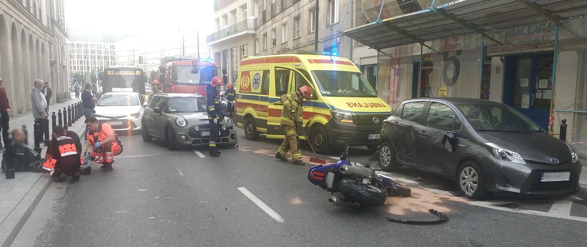 Na zdjęcie przedstawia sytuację zastaną przez strażaków po dojeździe na miejsce zdarzenia. Na zdjęciu z lewej strony na chodniku znajduje się poszkodowana osoba oraz ratownicy medyczni w pomarańczo-czarnych ubraniach którzy udzielają osobie pomocy medycznej. Po prawej stronie widać leżący niebieski motocykl oraz karetkę Pogotowia Ratunkowego z kolorze żółto-czerwono-niebieskim. Na zdjęciu widać tez strażaków w piaskowych oraz żółto-czarnych ubraniach ochronnych którzy zabezpieczają miejsce zdarzenia oraz zasypali sorbentem plamę oleju.