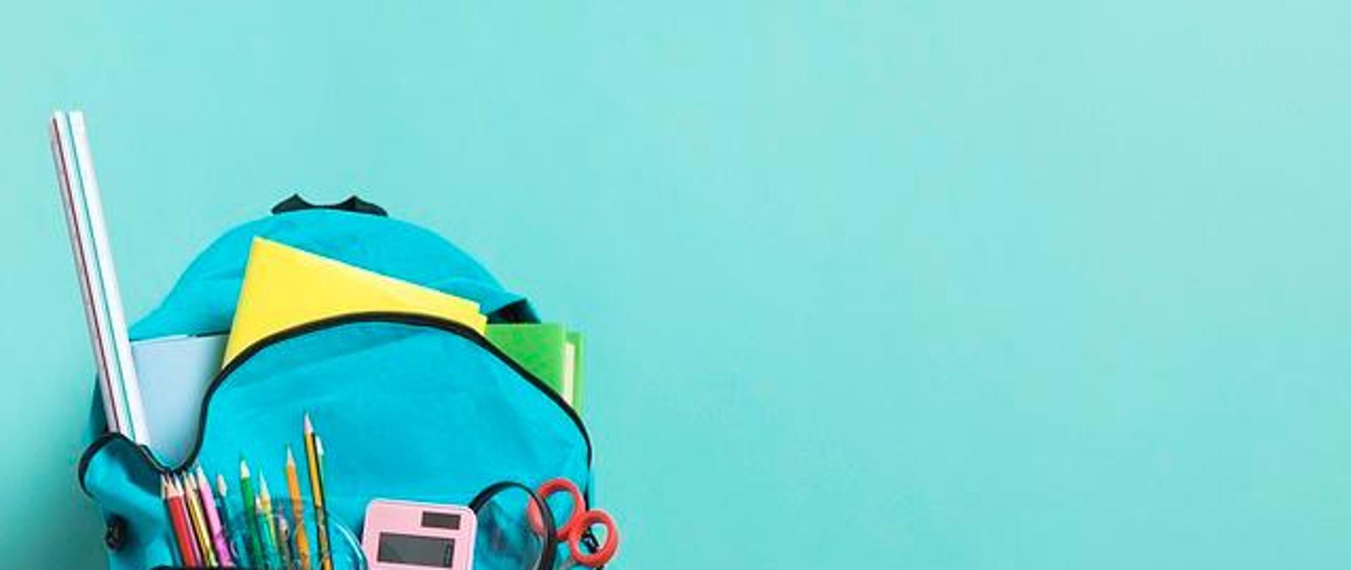 na zdjęciu widoczny niebieski plecak na jasnoniebieskim tle. obok plecaka zielone jabłko. z plecaka wystają kredki ołówkowe, kalkulator, lupa, nożyczki oraz książki