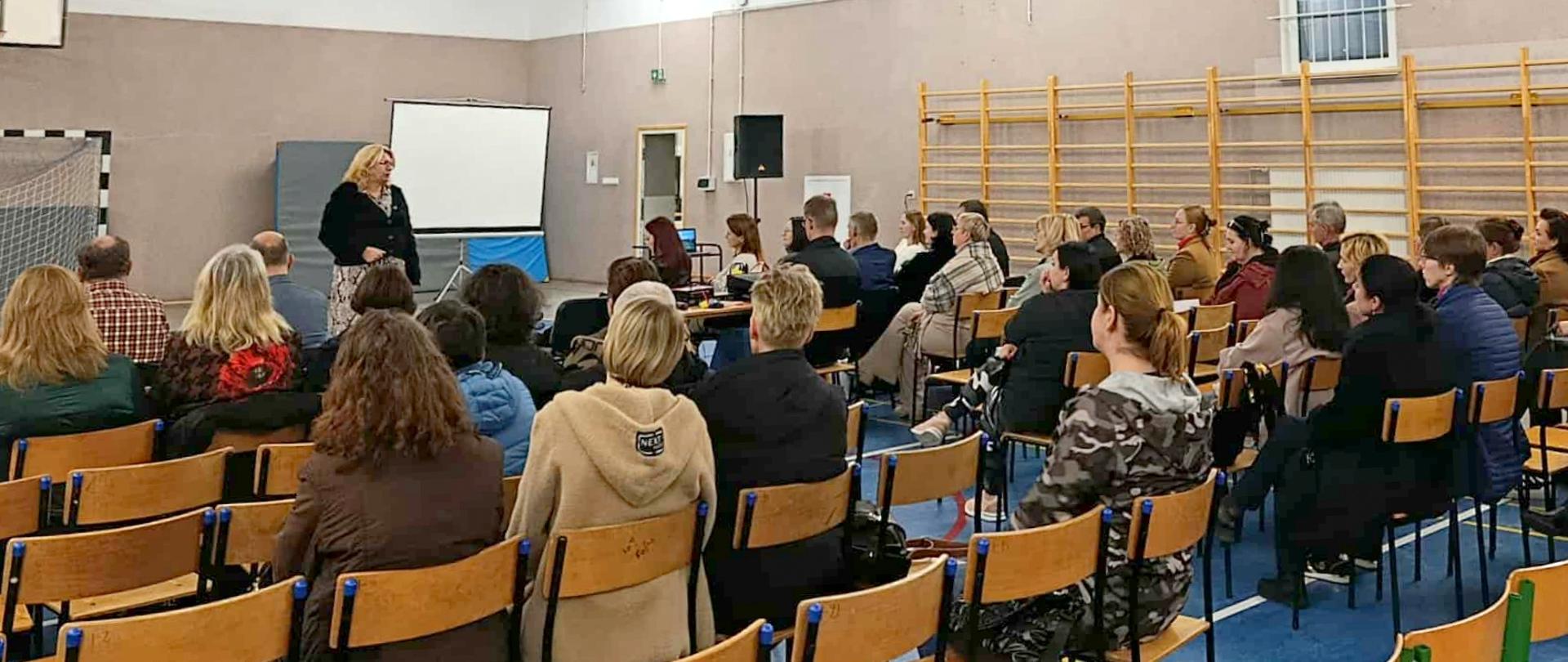 Kobieta prowadzi prelekcję, przed nią siedzi publiczność, słuchając wykładu.