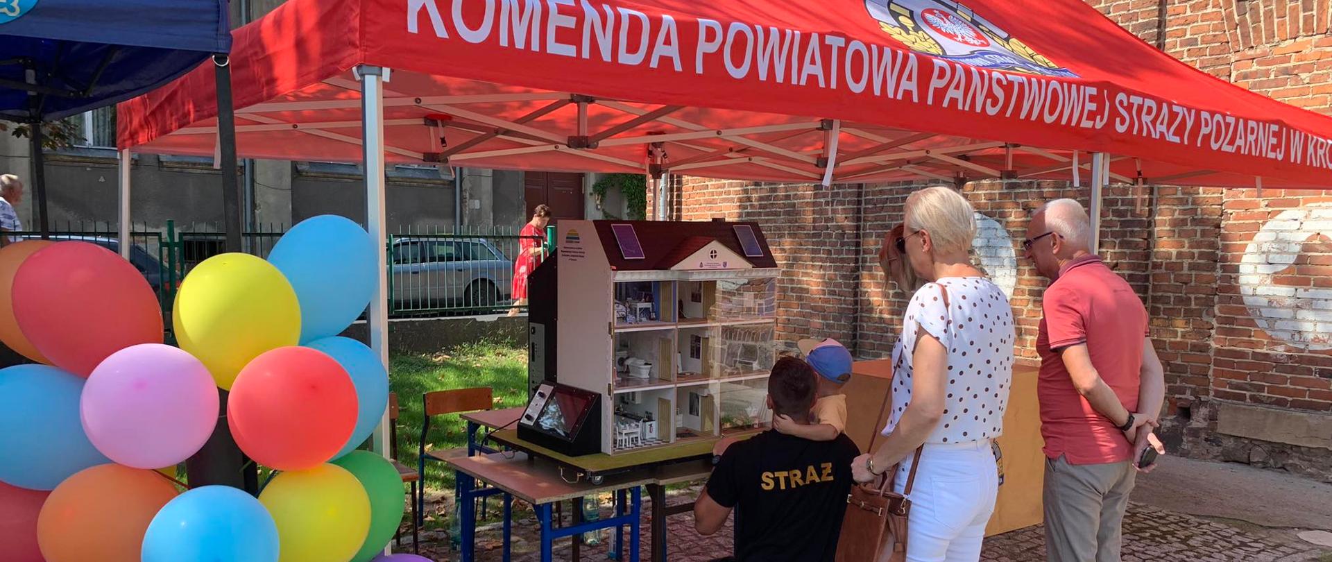 Zdjęcie przedstawia stoisko edukacyjne straży pożarnej. Pod namiotem stelażowym z czerwonym nakryciem znajduje się symulator zagrożeń w formie domku dla lalek w skali 1:12. Prelekcję prowadzą strażacy dla zebranych wokół namiotu uczestników dożynek