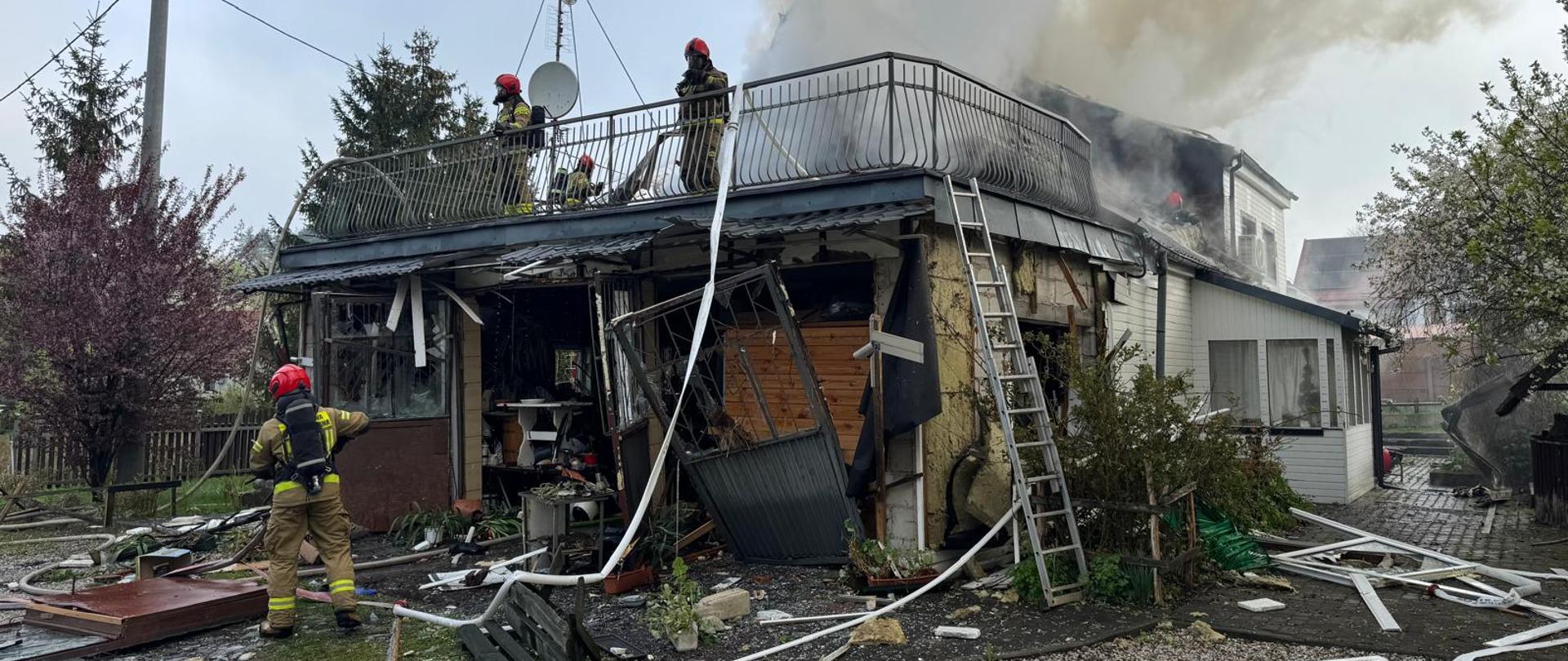 W centralnej części zdjęcia znajduje się budynek mieszkalny, częściowo zniszczony. Dookoła są porozrzucane materiały budowlane. Na dachy budynku i przed nim widoczne są sylwetki strażaków.