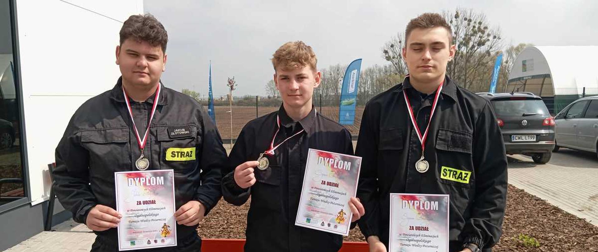 Zdjęcie przedstawia trzech młodych mężczyzn stojących obok siebie na zewnątrz, prawdopodobnie po zakończeniu zawodów strażackich. Wszyscy są ubrani w czarne mundury strażackie z charakterystycznymi żółtymi napisami „STRAŻ” na piersiach oraz odblaskowymi pasami na spodniach.
Oto szczegóły zdjęcia:
Postacie: Trzej młodzi strażacy stoją w rzędzie, patrząc w stronę obiektywu. Każdy z nich trzyma w dłoniach dyplom z napisem „DYPLOM ZA UDZIAŁ” i ma na szyi medal na biało-czerwonej wstążce.
Otoczenie: Mężczyźni stoją przed czerwoną ławką. W tle widać nowoczesny budynek o białych i szarych ścianach, fragment parkingu z samochodami oraz puste pole. Na dalszym planie widoczne są niebieskie flagi reklamowe i drzewa bez liści, co sugeruje wczesną wiosnę lub jesień.
Atmosfera: Scena ma charakter oficjalny i radosny, dokumentujący sukces lub uczestnictwo w wydarzeniu związanym ze strażą pożarną.