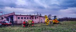 Na zdjęciu śmigłowiec lotniczego pogotowia ratunkowego i ratownicy transportujący osobę poszkodowaną na tle łąki i budynku z elewacją koloru różowego. W tle widoczne pojazdy straży pożarnej koloru czerwonego.