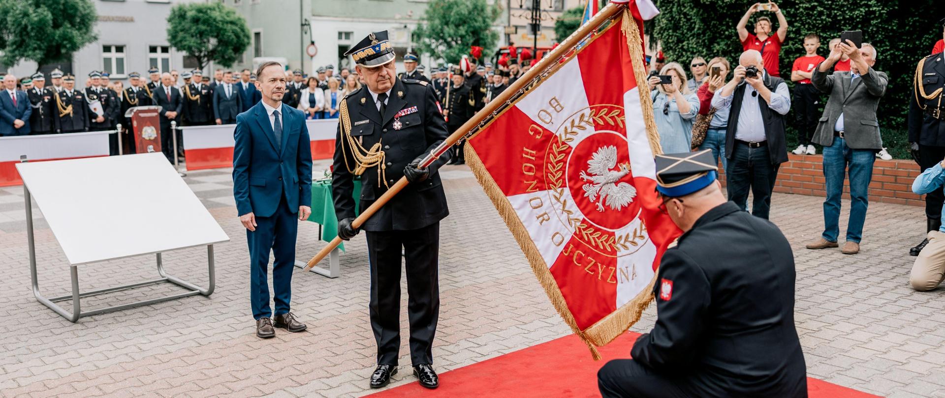 Zastępca komendanta głównego PSP trzyma pochylony sztandar. Przed sztandarem uklęknął komendant powiatowy PSP i całuje płat sztandaru.