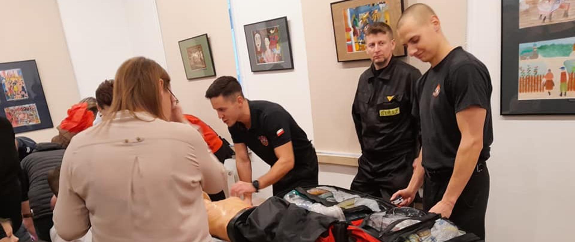 Na zdjęciu widać trzech strażaków w czarnych strojach, którzy pokazują zgromadzonym dzieciom i osobom dorosłym torbę medyczną oraz ćwiczenia na fantomie. Na ścianach wiszą obrazy.