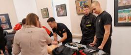 Na zdjęciu widać trzech strażaków w czarnych strojach, którzy pokazują zgromadzonym dzieciom i osobom dorosłym torbę medyczną oraz ćwiczenia na fantomie. Na ścianach wiszą obrazy.