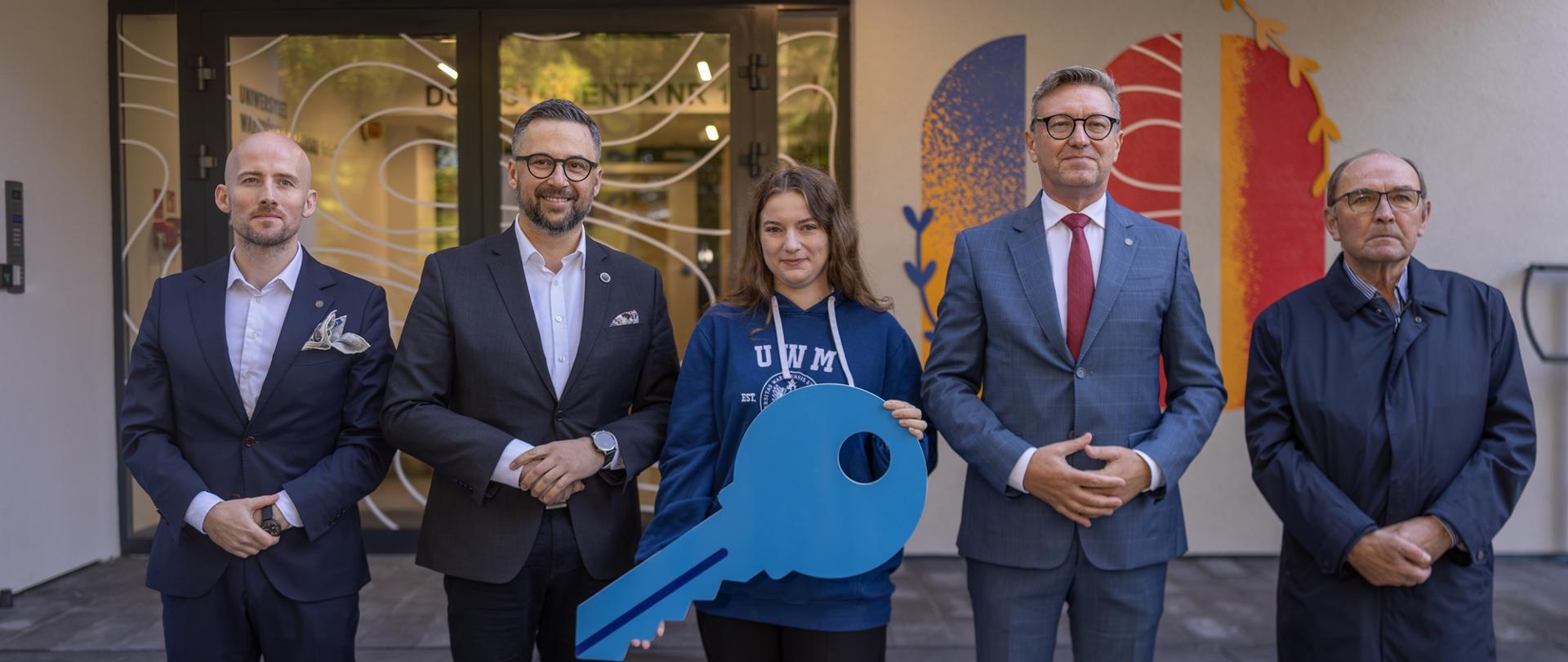 Przed wejściem do budynku stoi pięć osób, od lewej mężczyzna w granatowym garniturze, minister Kulasek, młoda dziewczyna w granatowiej bluzie z napisem UWM trzymająca wielki niebieski symboliczny klucz i dwóch mężczyzn w garniturach.