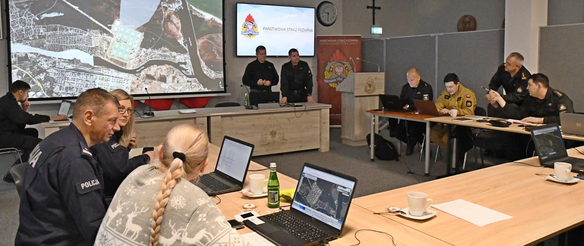 Dwóch strażaków stoi za stołem za nimi jest rozłożony ekran multimedialny z mapą przedstawiciele służb oraz instytucji siedzą za stołami przed laptopami podczas ćwiczenia w formie sztabowej.