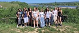 Grupa uczniów stojąca na wzgórzu z widokiem na rzekę i zielone tereny podczas wyjazdu studyjnego.