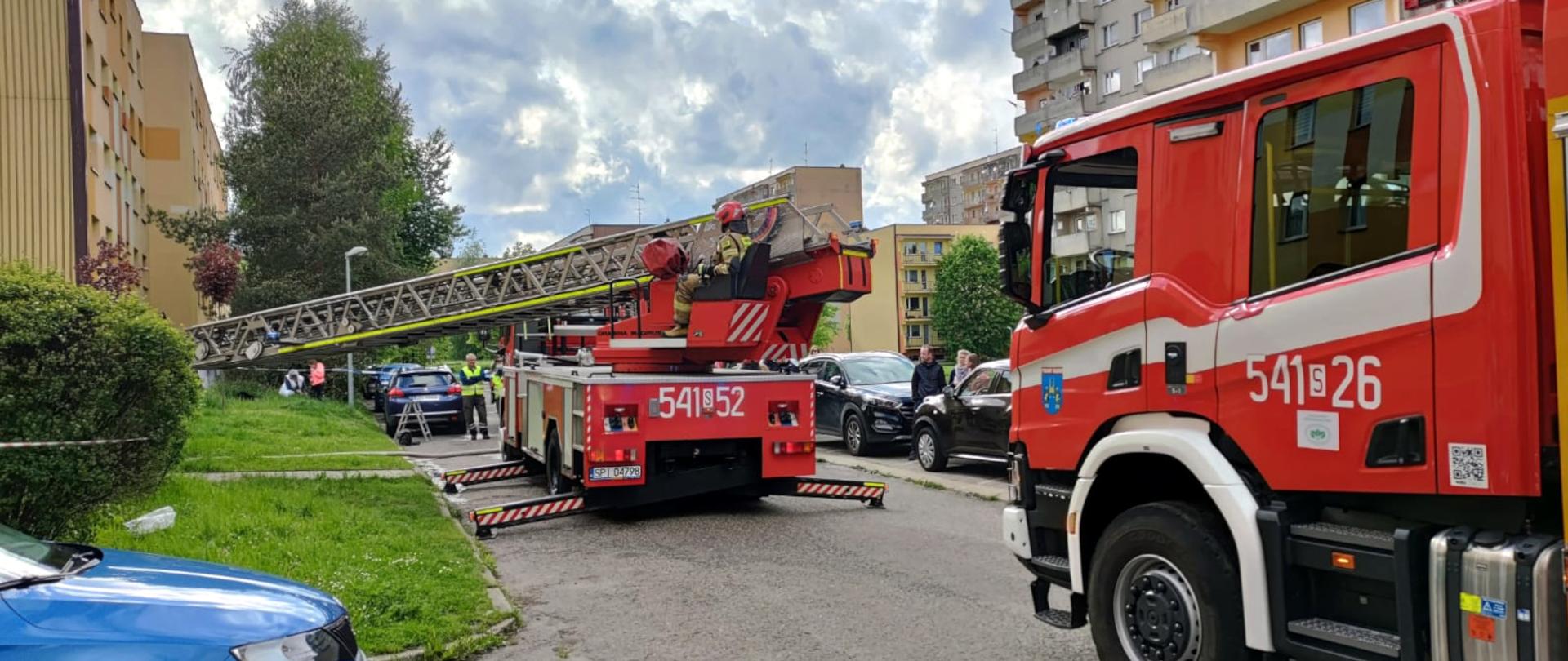 Zdjęcie przedstawia samochód specjalny drabinę pożarniczą. W pojeździe na stanowisku operatora drabiny siedzi strażak w ubraniu specjalnym. Obok stoi ciężki samochód ratowniczo-gaśniczy. Obydwa samochody stoją na ulicy, na osiedlu mieszkaniowym pośród bloków mieszkalnych.