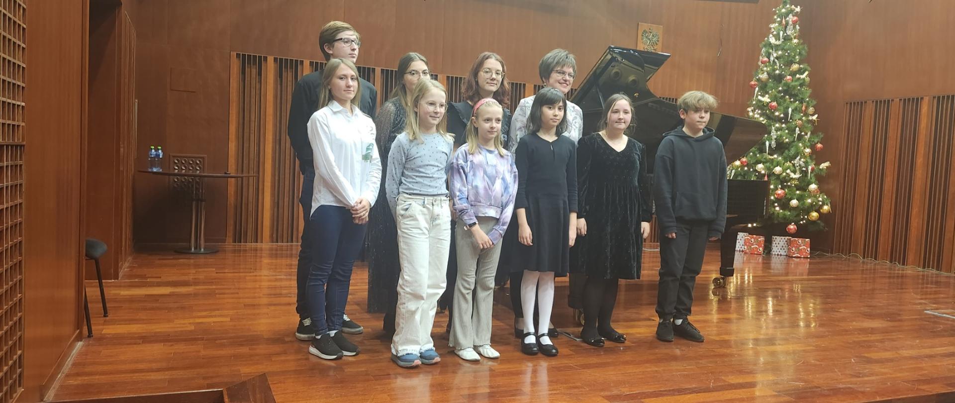 Zdjęcie uczestników warsztatów pianistycznych wraz z prof. Oksaną Rapitą w sali koncertowej szkoły. W tle brązowa scena, fortepian oraz choinka.