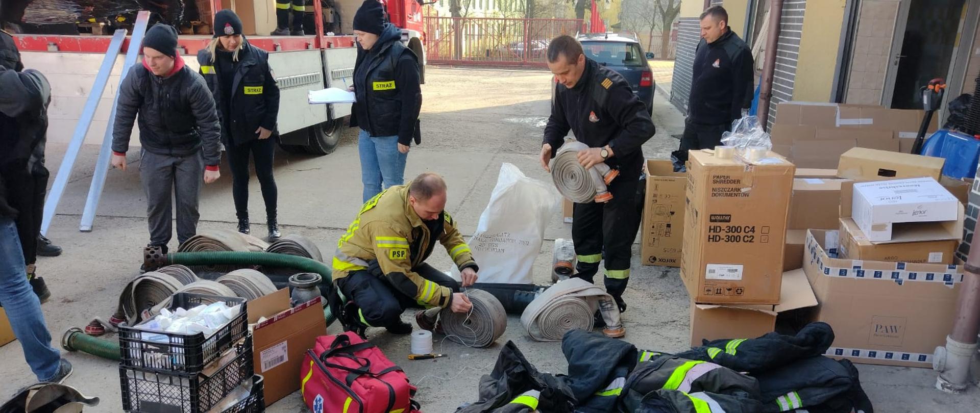 Dzień. Z lewej strony widać samochód ciężarowy oplandekowany plandeką w kolorze czerwonym, kabina pojazdu czerwona. Plandeka zarzucona do góry, skrzynia pojazdu częściowo wypełniona pakunkami i pudłami. Z prawej strony zdjęcia widać mur i częściowo podniesioną do góry bramą wjazdową w kolorze czerwonym. Pomiędzy pojazdem a ścianą na ziemi leży dużo rożnego rodzaju sprzętu ppoż. Pomiędzy sprzętem znajdują się strażacy i osoby cywilne pakujące sprzęt do pudeł i worków.