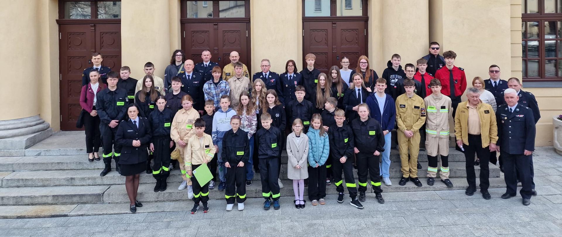 Duża grupa dzieci, młodzieży i dorosłych stoi na schodach przed reprezentacyjnym budynkiem z kolumnami i wysokimi drzwiami. Wśród uczestników widoczne są osoby w mundurach strażackich oraz młodzież w strojach z elementami odblaskowymi. Wszyscy pozują do wspólnego zdjęcia, co sugeruje zakończenie wydarzenia lub konkursu związanego ze strażą pożarną.