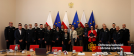 Zdjęcie zbiorowe - kilkadziesiąt osób - funkcjonariuszy PSP i cywili , stoją w grupie, za nimi rząd flag - naprzemiennie biało-czerwone i unijne. Nad nimi godło - przeł w kolorze złotym. Przed nimi widoczne rzędy stołów...