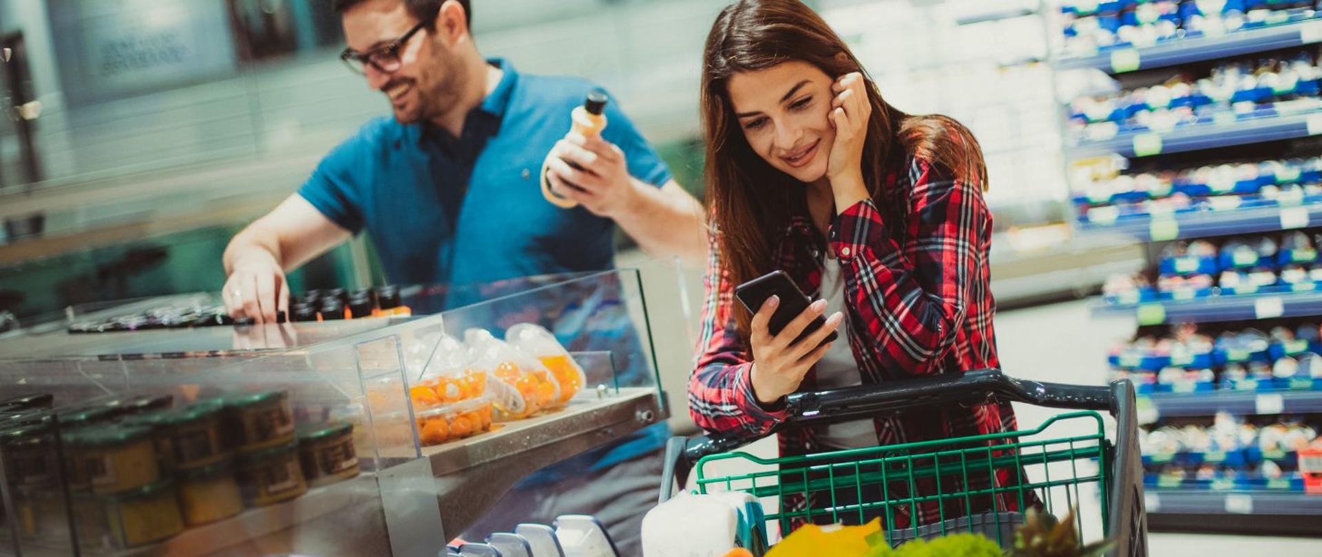 na zdjęciu widnieją kobieta i mężczyzna robiący zakupy w sklepie. Kobieta patrzy w telefon, opierając się na koszyku na zakupy. Mężczyzna wybiera soki w butelkach plastikowych. 