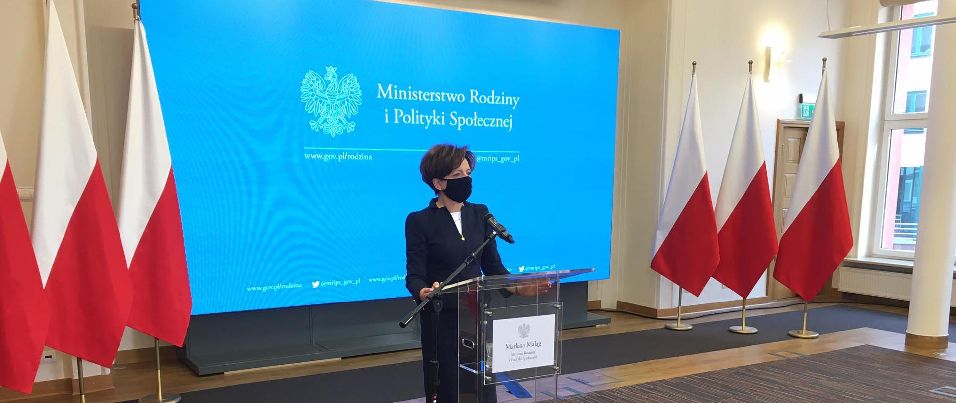 Sala Ministerstwa Rodziny i Polityki Społecznej. Minister Maląg informuje, stojąc przy mównicy o Solidarnościowym Korpusie Wsparcia Seniorów. 