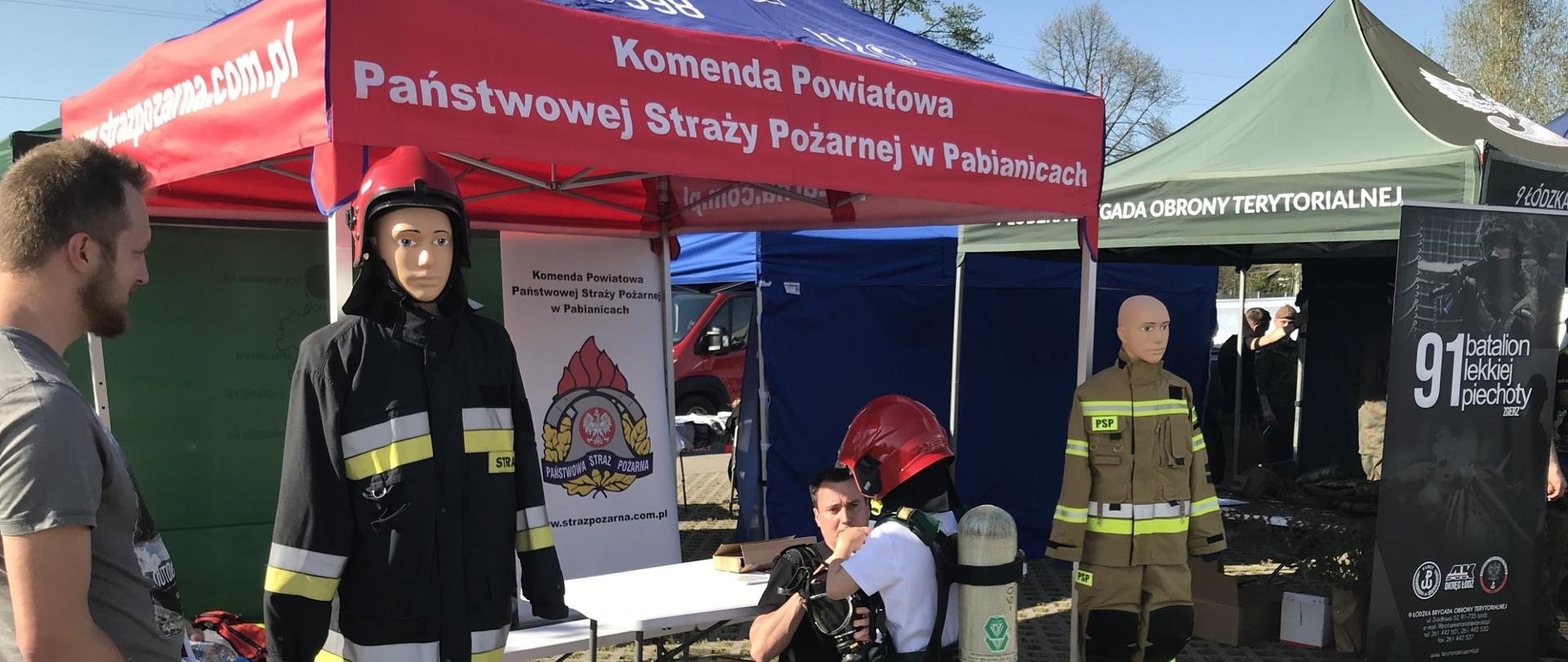 Na zdjęciu przy stoisku KP PSP w Pabianicach dziecko przymierza hełm strażacki oraz zakłada na plecy aparat powietrzny z pomocą strażaka. W tle widoczny namiot Wojsk Obrony Terytorialnej.