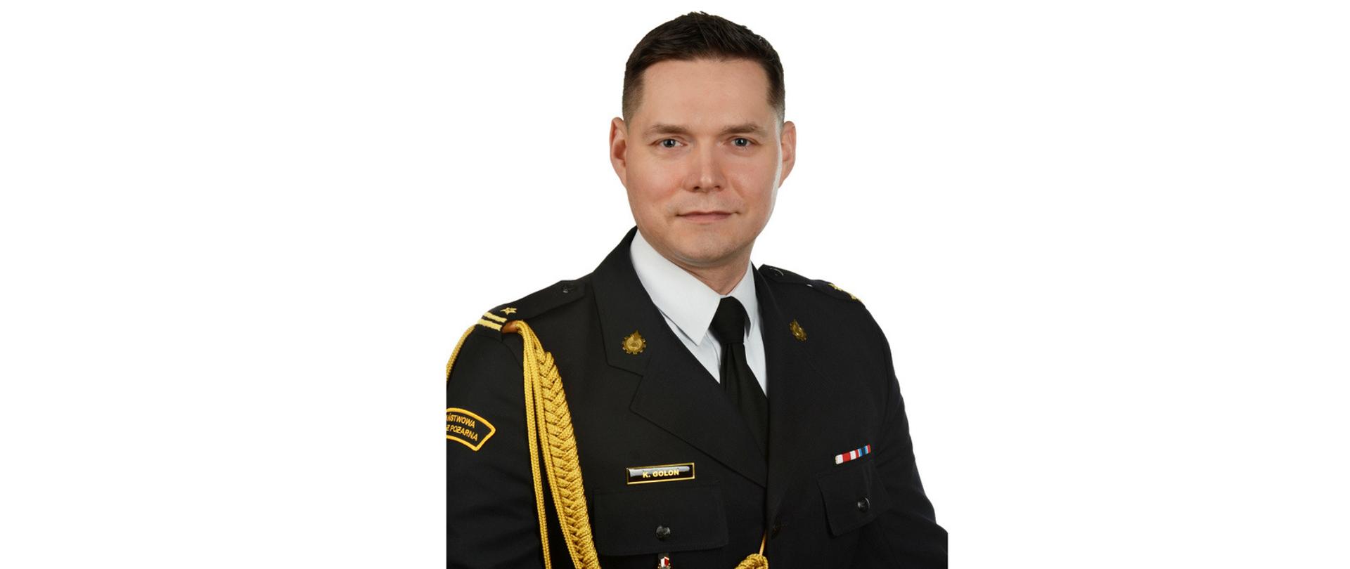 Funkcjonariusz w czarnym mundurze galowym ze złotym sznurem. na lewym ramieniu napis Państwowa Straż Pożarna. Na klapach gabardyny zawieszone różne odznaczenia.