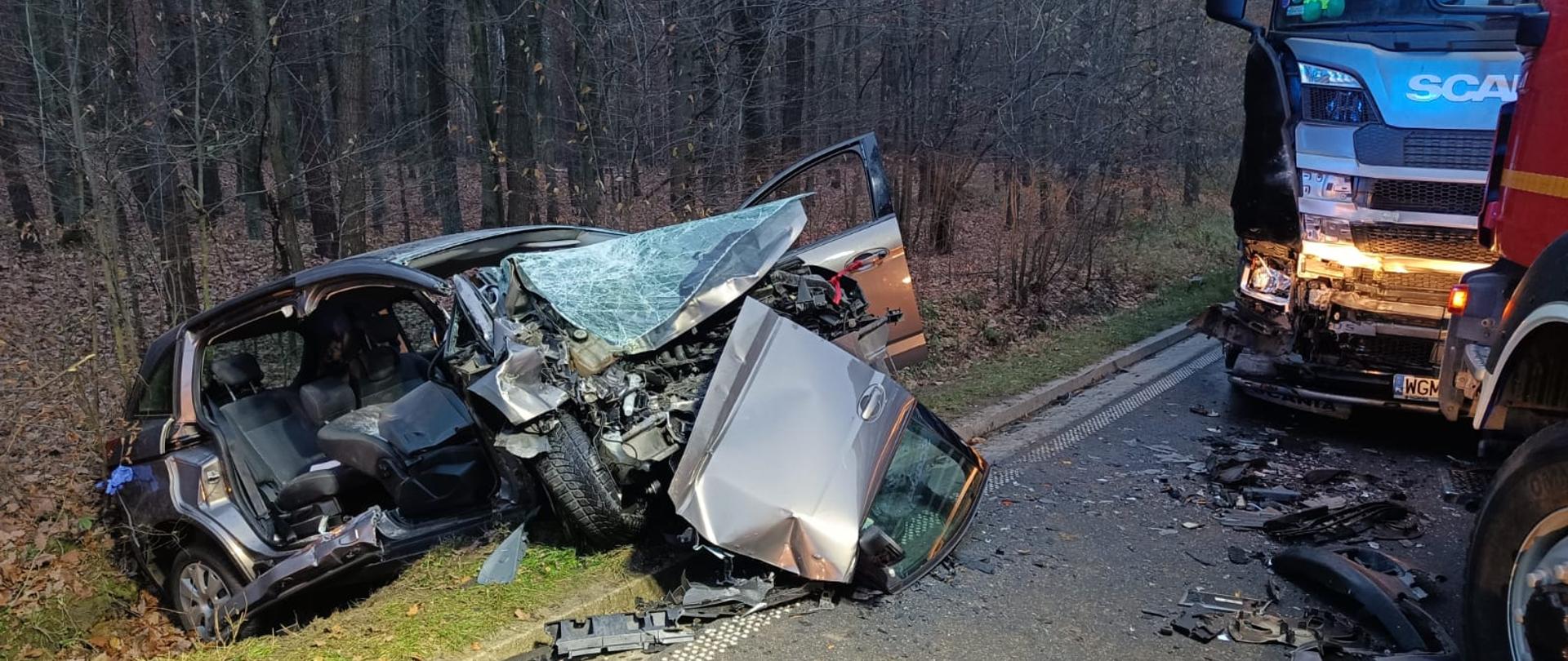 Uszkodzony ciągnik siodłowy stoi na ulicy. W rowie stoi samochód osobowy z bardzo zniszczonym przodem.