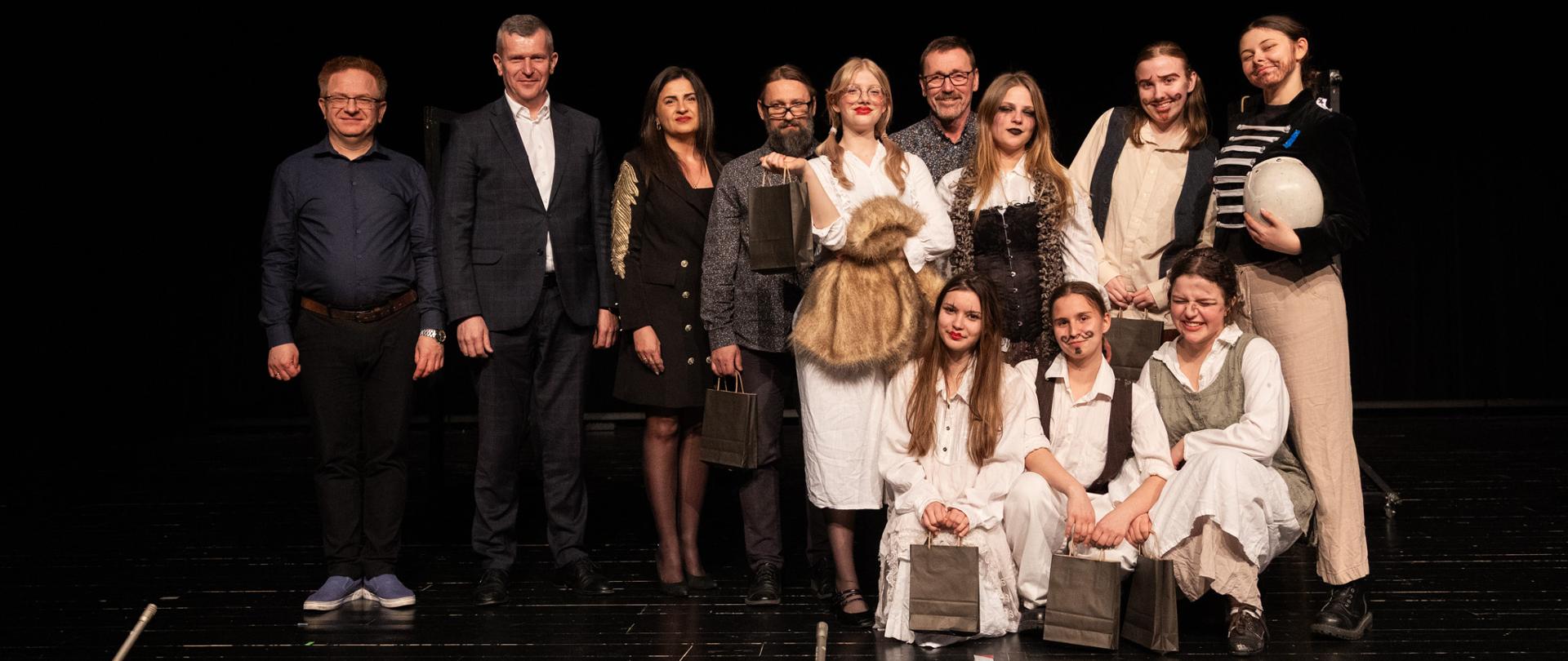 Na scenie do zdjęcia pozuje grupa osób: po prawej stronie stoi elegancko ubranych dwóch mężczyzn i kobieta, a z prawej strony jest siedem dziewcząt w kostiumach teatralnych, za nimi dwóch mężczyzn, trzymają w rękach czarne papierowe torebki.